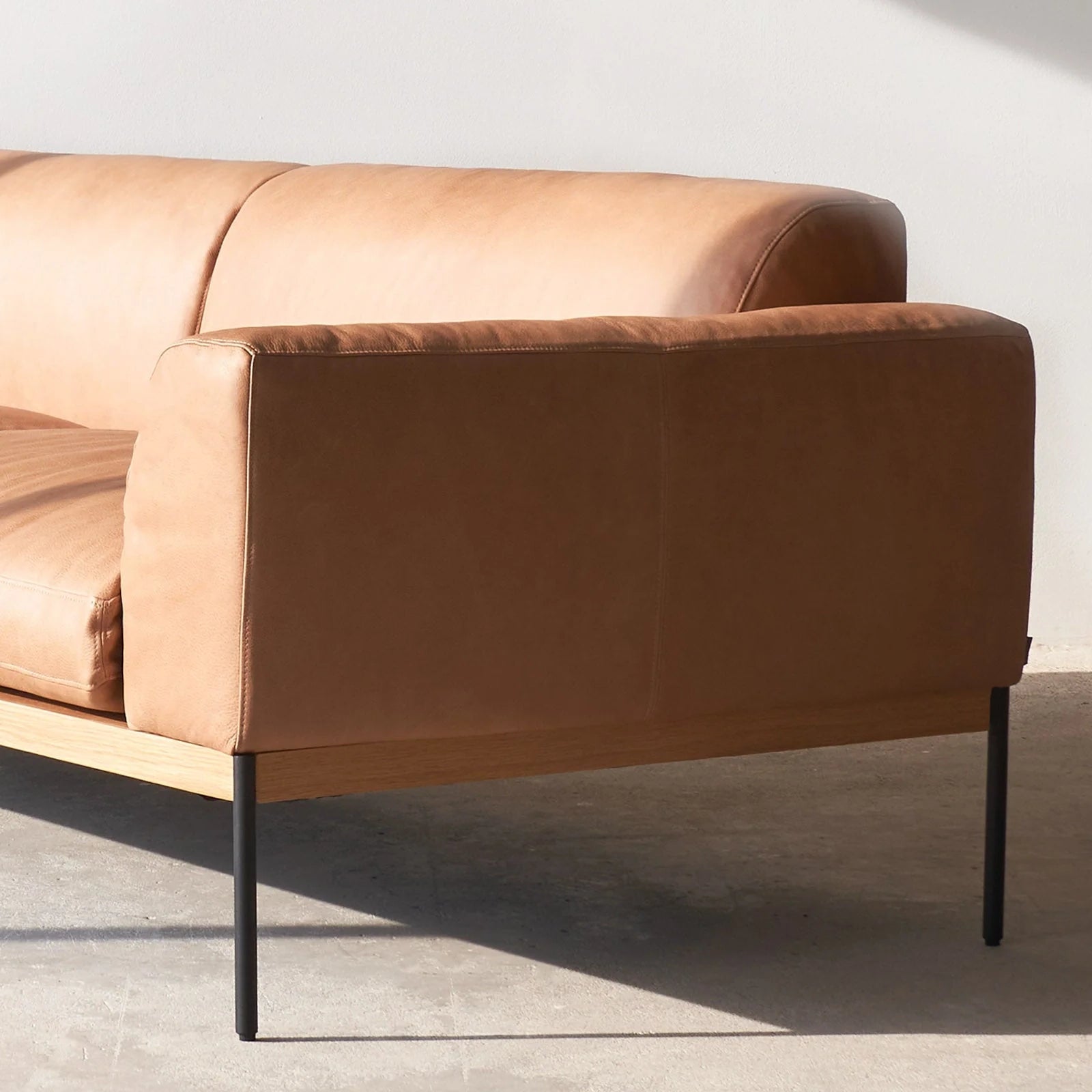 The Wooden Stories Department Modular Sofa | Leather • M4 Left features modern tan leather upholstery, an FSC-certified oak base, and slim black metal legs, shown here on a concrete floor with sunlight casting shadows behind it.
