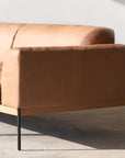 The Wooden Stories Department Modular Sofa | Leather • M4 Left features modern tan leather upholstery, an FSC-certified oak base, and slim black metal legs, shown here on a concrete floor with sunlight casting shadows behind it.
