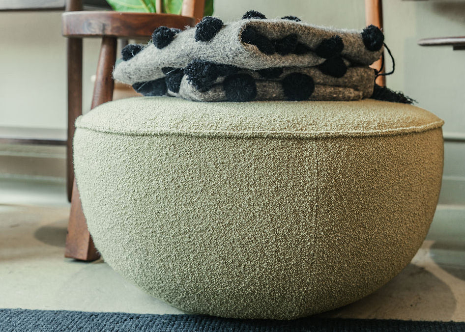 A Fable Ottoman | Fabric by Wooden Stories sits on a rug beside a folded gray and black blanket. Behind it, clean Danish lines appear in a wooden ladder holding another blanket, with a leafy plant, chair, and shelves with décor.