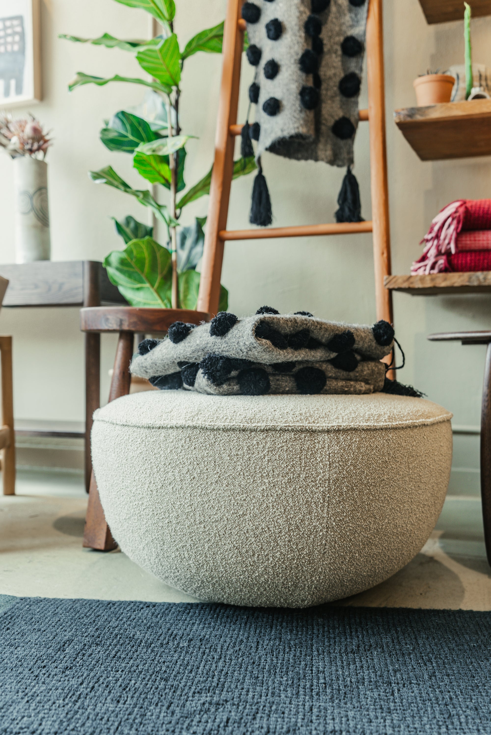 Round beige Fable ottoman with a patterned throw on top in a room with a wooden ladder and plants.