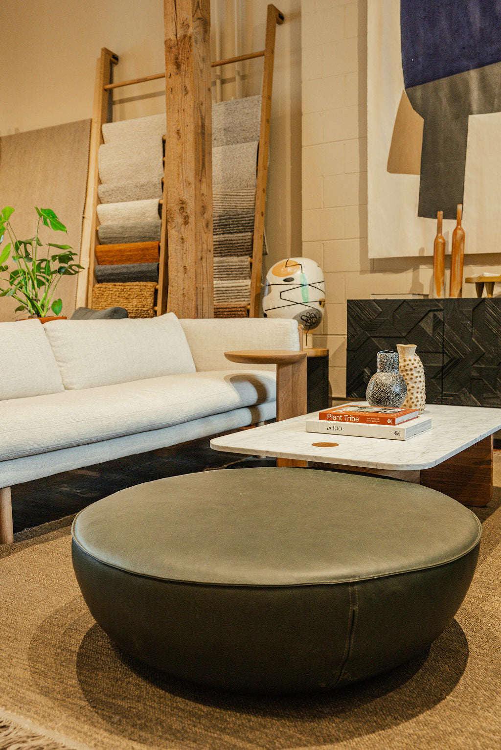 A modern living room with a light gray sofa, the Wooden Stories Fable Ottoman in leather as extra seating, a white marble coffee table displaying books and vases, textiles on wooden racks, a potted plant, and contemporary wall art.