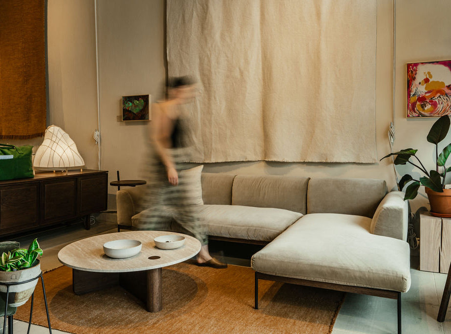 A blurred figure passes by the Wooden Stories Department Modular | Fabric • M2 Right sectional sofa in a cozy living room with earthy tones, abstract art, potted plants, a solid wood plinth coffee table, and soft ambient lighting.