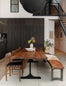 A modern dining area showcases the SOBU Leviathan Live Edge Dining Table in smoked acacia with black T-legs, paired with matching chairs and a bench topped with a fur throw. Dark cabinets, greenery, and a centerpiece warm the stylish space.