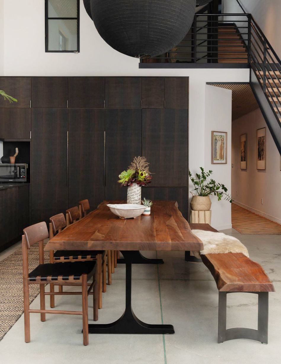 A modern dining area showcases the SOBU Leviathan Live Edge Dining Table in smoked acacia with black T-legs, paired with matching chairs and a bench topped with a fur throw. Dark cabinets, greenery, and a centerpiece warm the stylish space.