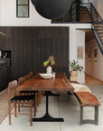 A modern dining area showcases the SOBU Leviathan Live Edge Dining Table in smoked acacia with black T-legs, paired with matching chairs and a bench topped with a fur throw. Dark cabinets, greenery, and a centerpiece warm the stylish space.