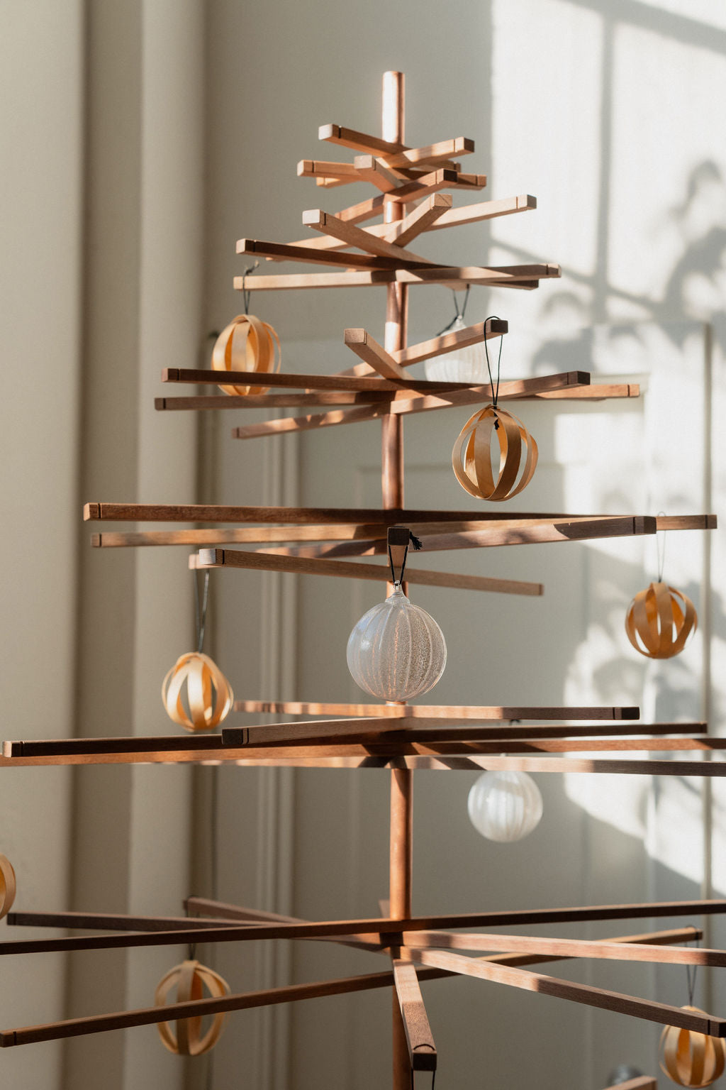 The Immortal Christmas Tree by SOBU is a minimalist, sustainable piece crafted from horizontal branches and decorated with simple white and gold ornaments. Sunlight creates soft shadows on the wall behind it.