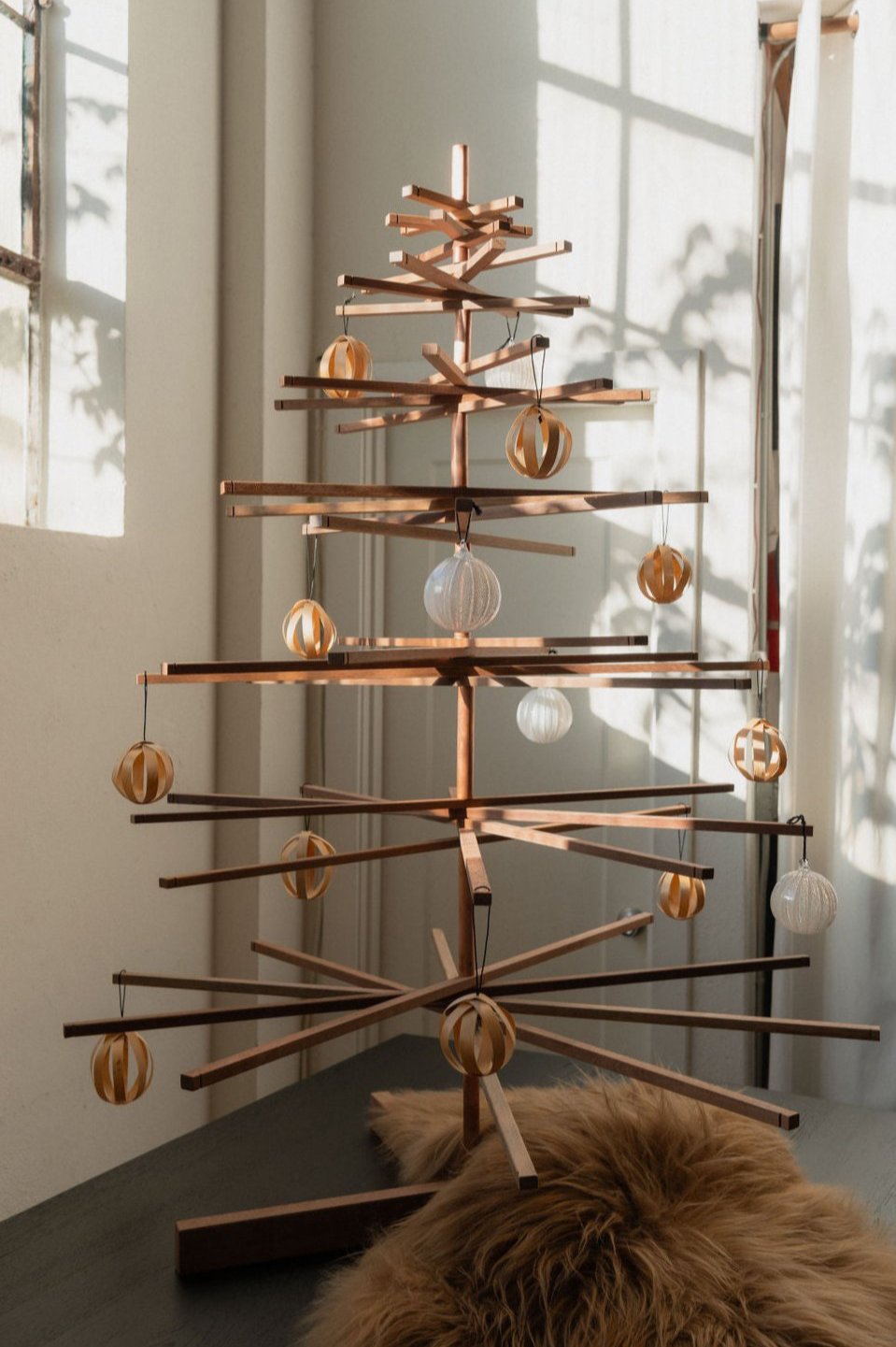 The SOBU Immortal Christmas Tree with geometric branches stands by a sunlit window, adorned with gold and white ornaments. A cozy brown fur rug rests at its base, creating a minimalist and sustainable holiday scene.