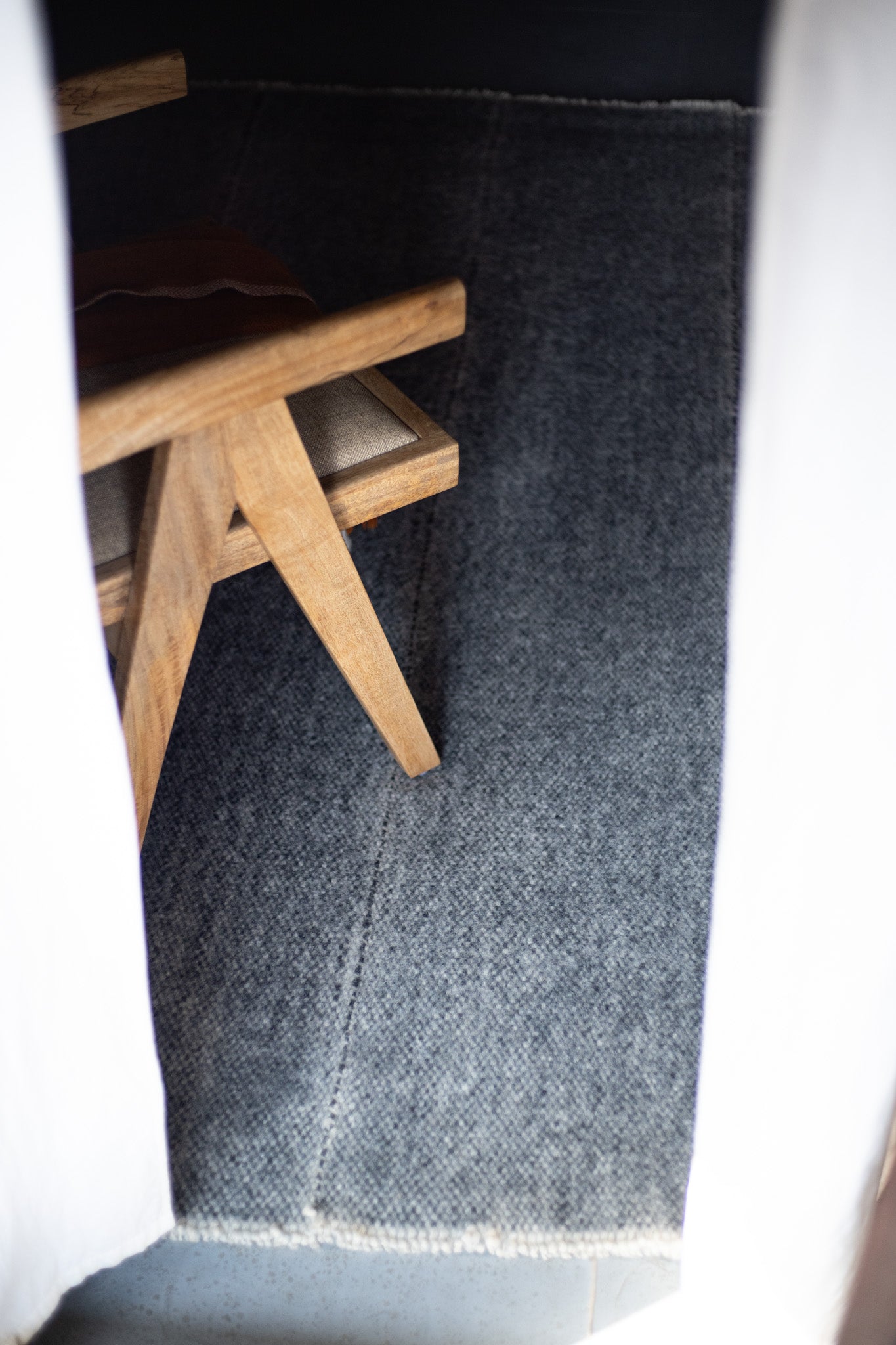 A wooden chair with beige cushioning is partially visible behind white curtains, set on a textured gray Radhi Rug - Silver by HIMALAYAN HERMITAGE.