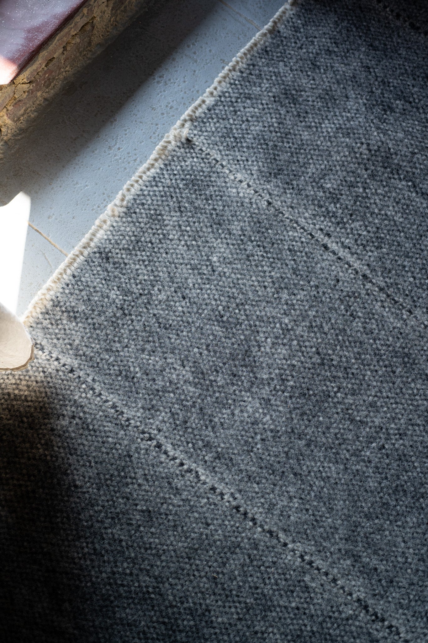A close-up of the HIMALAYAN HERMITAGE Radhi Rug - Silver shows its textured Nepali wool on a light floor, sunlight highlighting the hand-woven edge near a window while shadows fall across the rest.