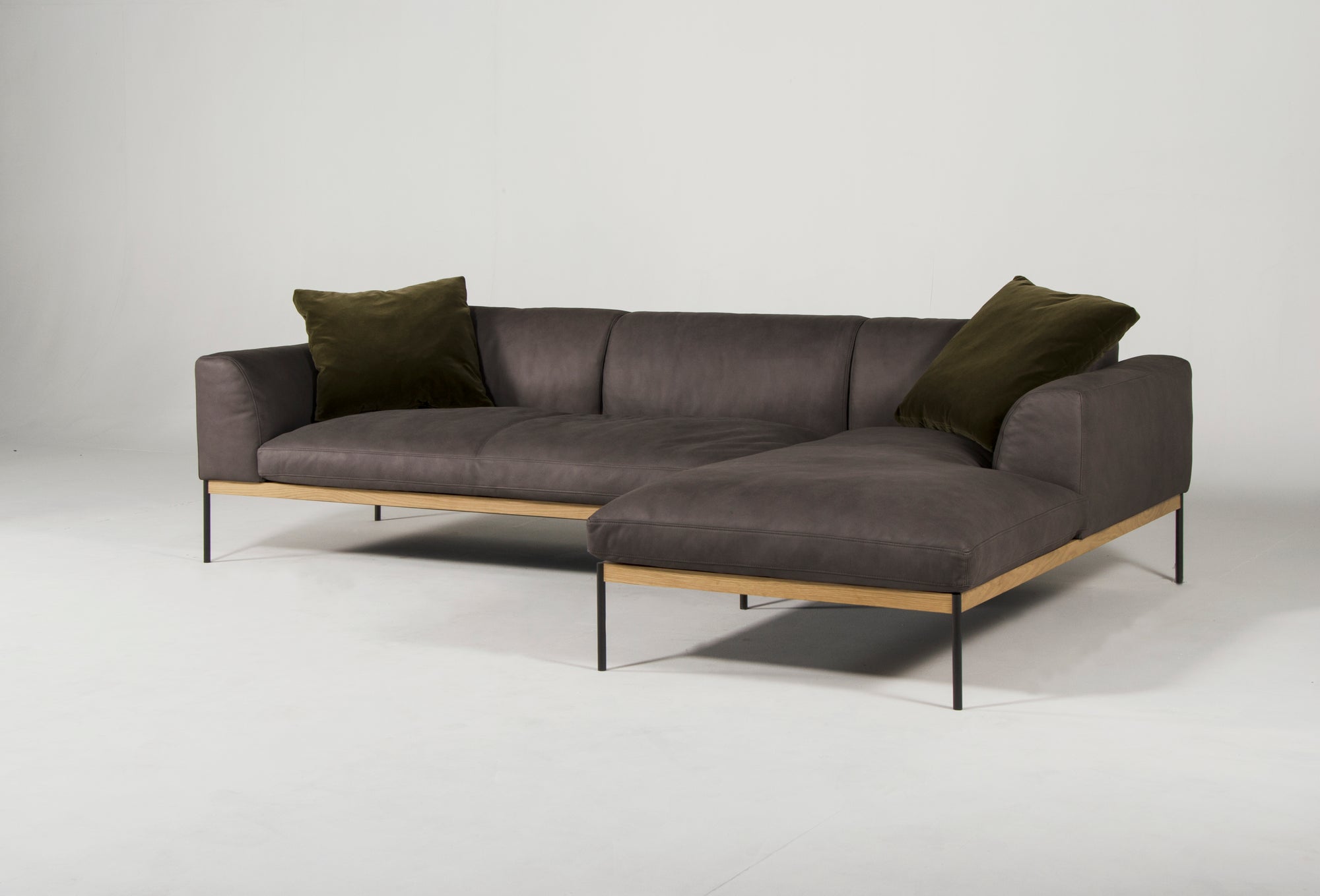 The Wooden Stories Department Modular Sofa | Leather • M2 Right features dark gray leather, a solid wood base, slim black metal legs, and two olive green pillows, displayed against a plain white background.