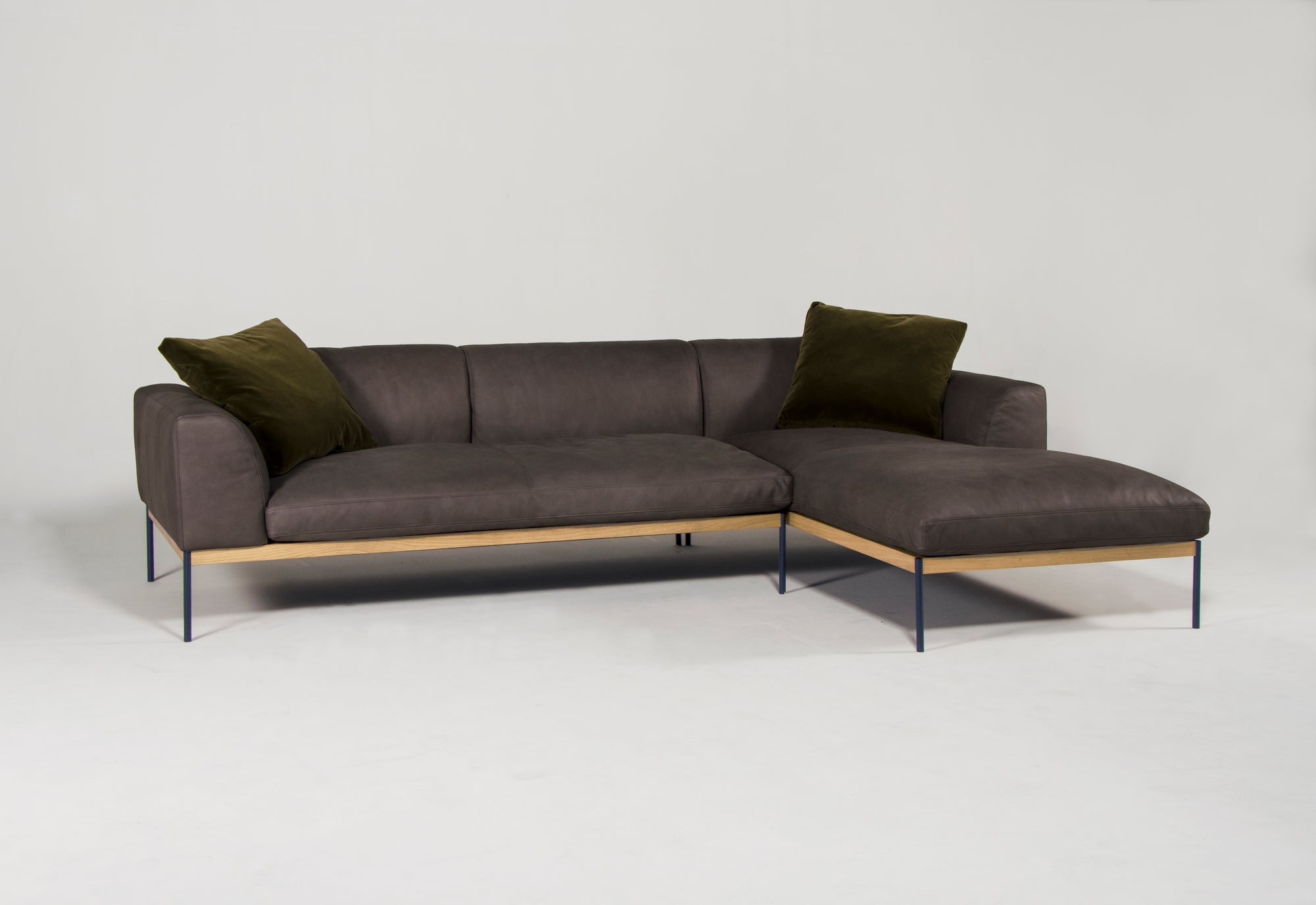 The Department Modular Sofa | Leather • M2 Right from Wooden Stories features brown leather upholstery, an FSC-certified American oak base, slim metal legs, and two dark green pillows, displayed against a plain white background.