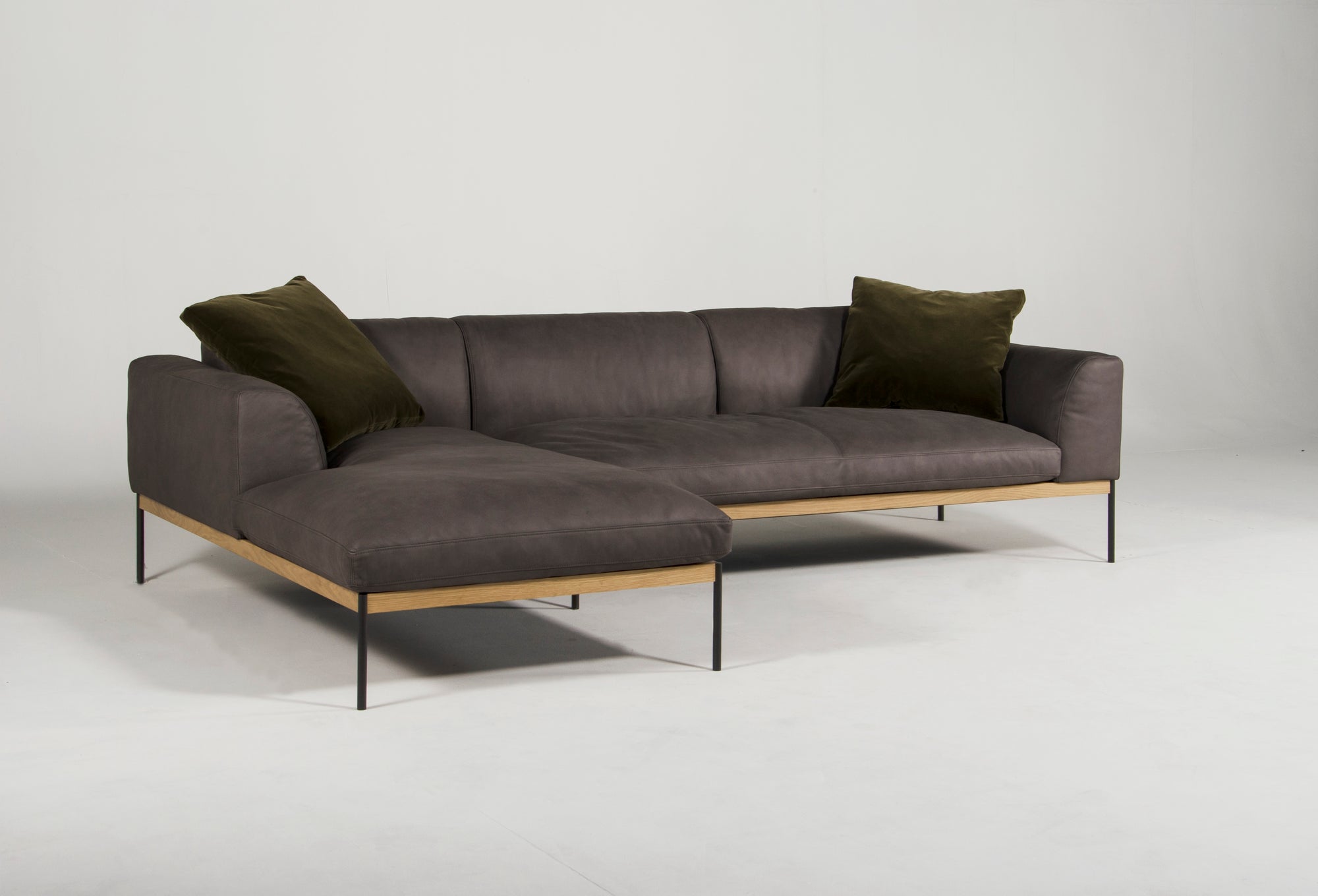 The Wooden Stories Department Modular Sofa | Leather • M2 Left features dark gray leather upholstery, an FSC-certified oak wood base, slim black metal legs, and two dark green throw pillows shown against a plain white background.