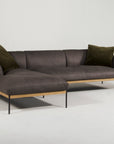 The Wooden Stories Department Modular Sofa | Leather • M2 Left features dark gray leather upholstery, an FSC-certified oak wood base, slim black metal legs, and two dark green throw pillows shown against a plain white background.