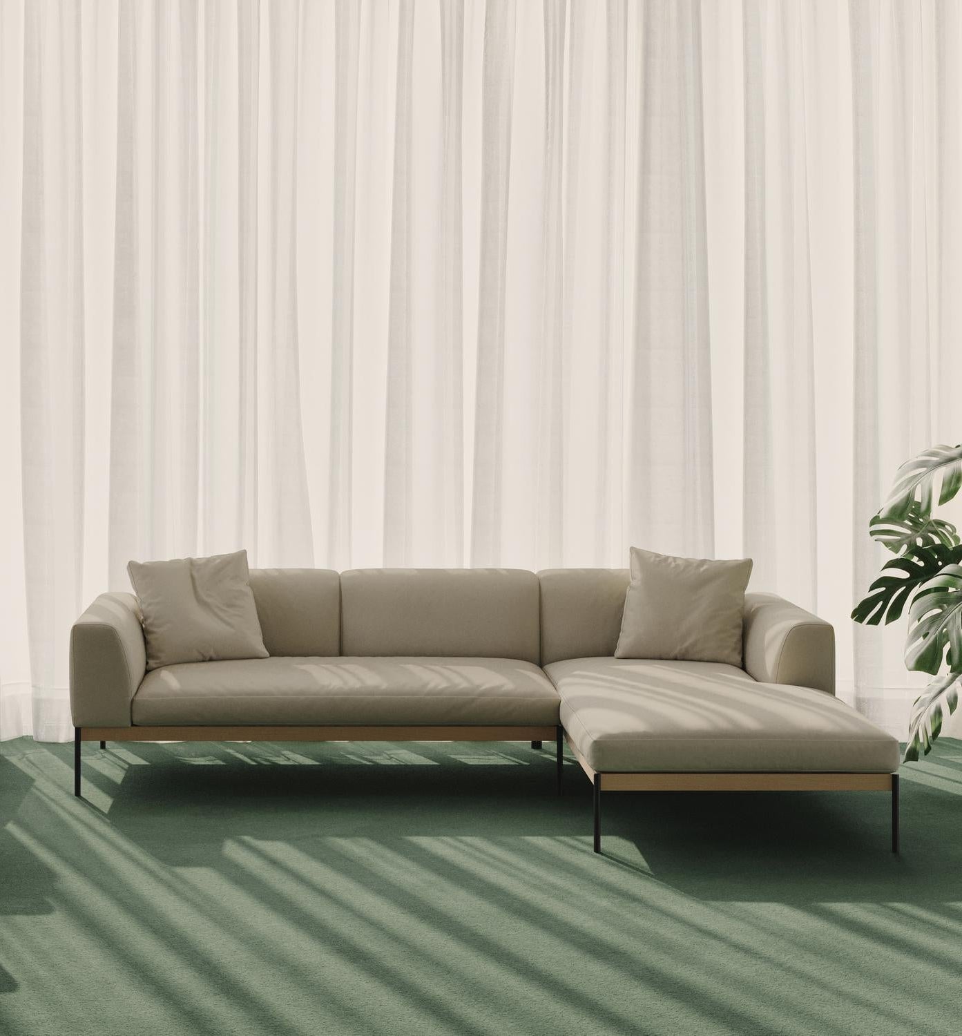 The Department Modular Sofa | Fabric • M2 Right by Wooden Stories in beige sits on a green carpet before sheer white curtains, sunlight casting shadows nearby, while a leafy potted plant stands beside its solid wood Danish-inspired base.