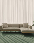 The Department Modular Sofa | Fabric • M2 Right by Wooden Stories in beige sits on a green carpet before sheer white curtains, sunlight casting shadows nearby, while a leafy potted plant stands beside its solid wood Danish-inspired base.