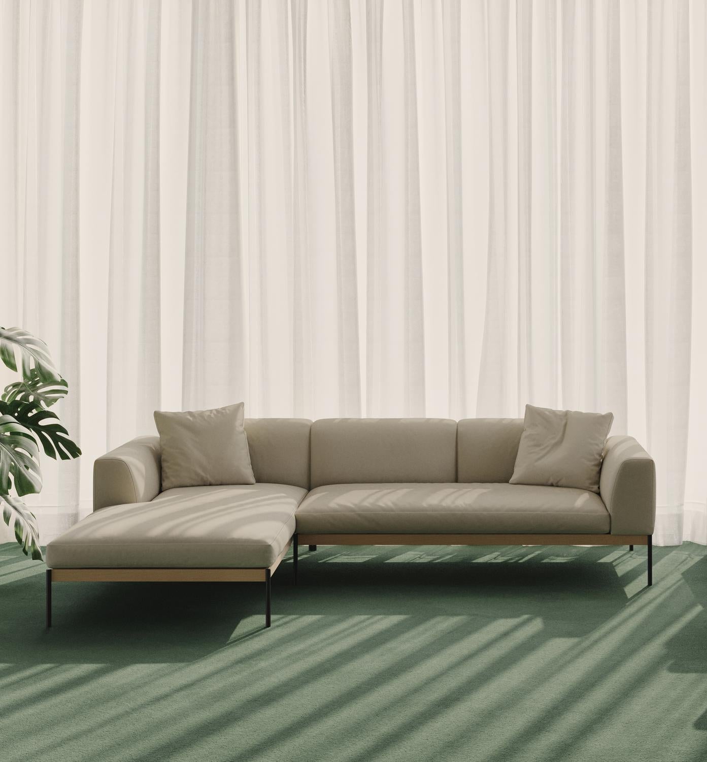 The Wooden Stories Department Modular Sofa | Fabric • M2 Left in beige, with two cushions, rests on a green carpet near sheer white curtains. Sunlight filters in, casting soft shadows, while a leafy plant stands beside its solid wood base.