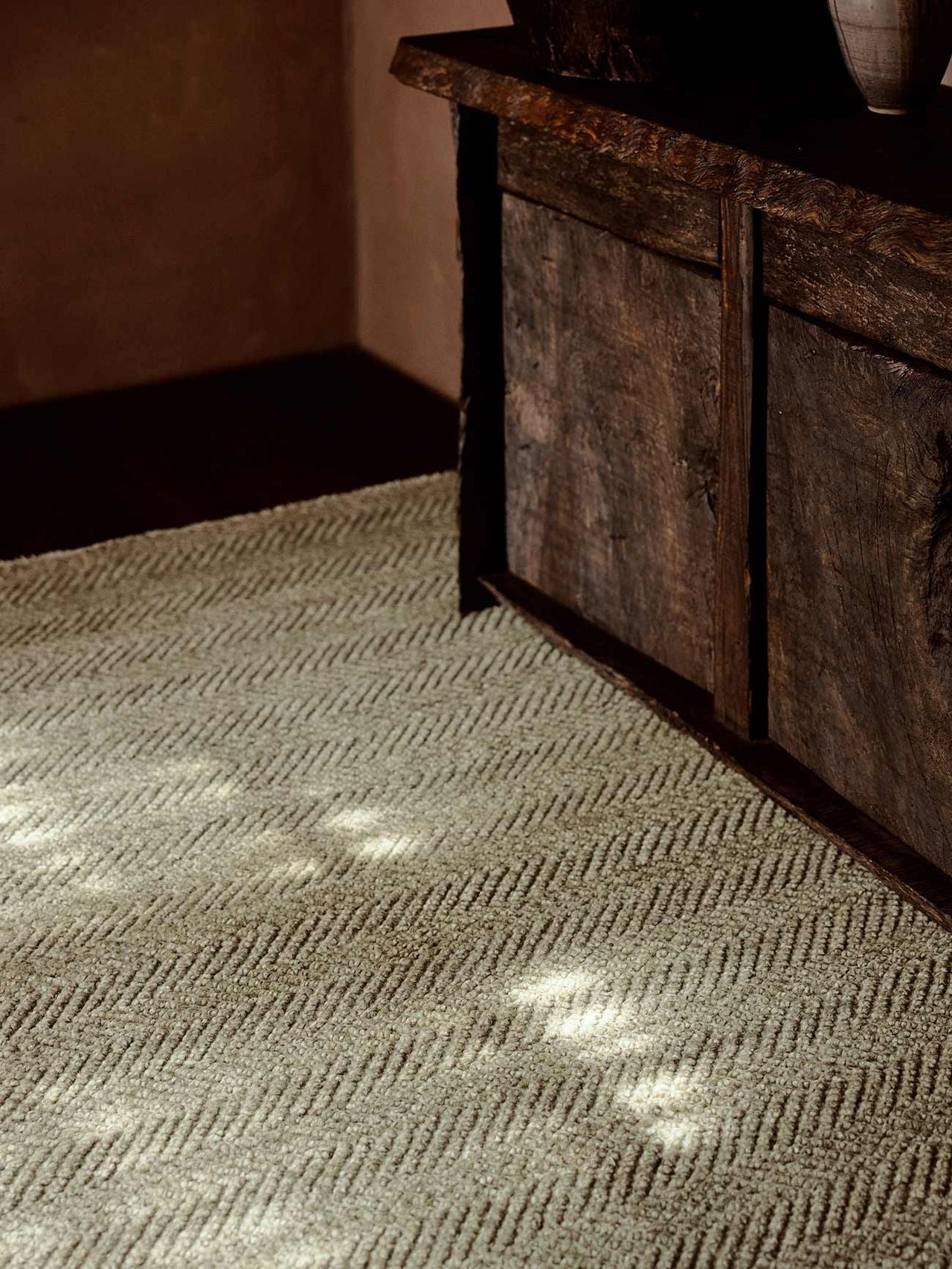 Sunlight streams onto a cream-colored Armadillo&Co Crest Rug - Mica with a subtle chevron pattern, positioned next to a rustic wooden cabinet with dark, weathered doors.