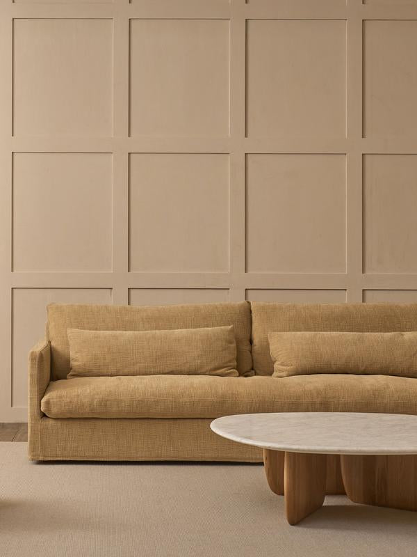 A beige couch with matching cushions sits before a paneled wall, next to the Wooden Stories Eden Coffee Table with a stone top, creating a minimalist living room infused with neutral tones and Scandinavian-inspired simplicity.