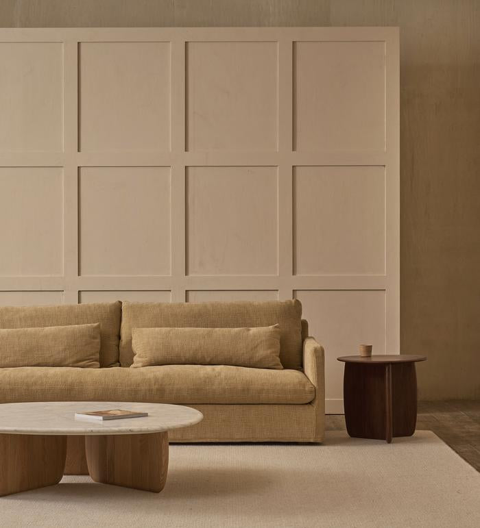 A minimalist living room with a beige sofa, two matching cushions, the Wooden Stories Eden Coffee Table with a stone top and book, an American oak side table with a cup, and a textured wall featuring rectangular paneling.