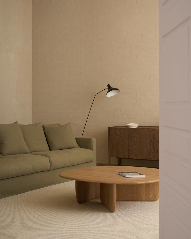 A calm, uncluttered living room features a green sofa, floor lamp, neutral walls and carpet, brown sideboard, and the Wooden Stories Eden Coffee Table in Light Oak with a magazine resting on top.