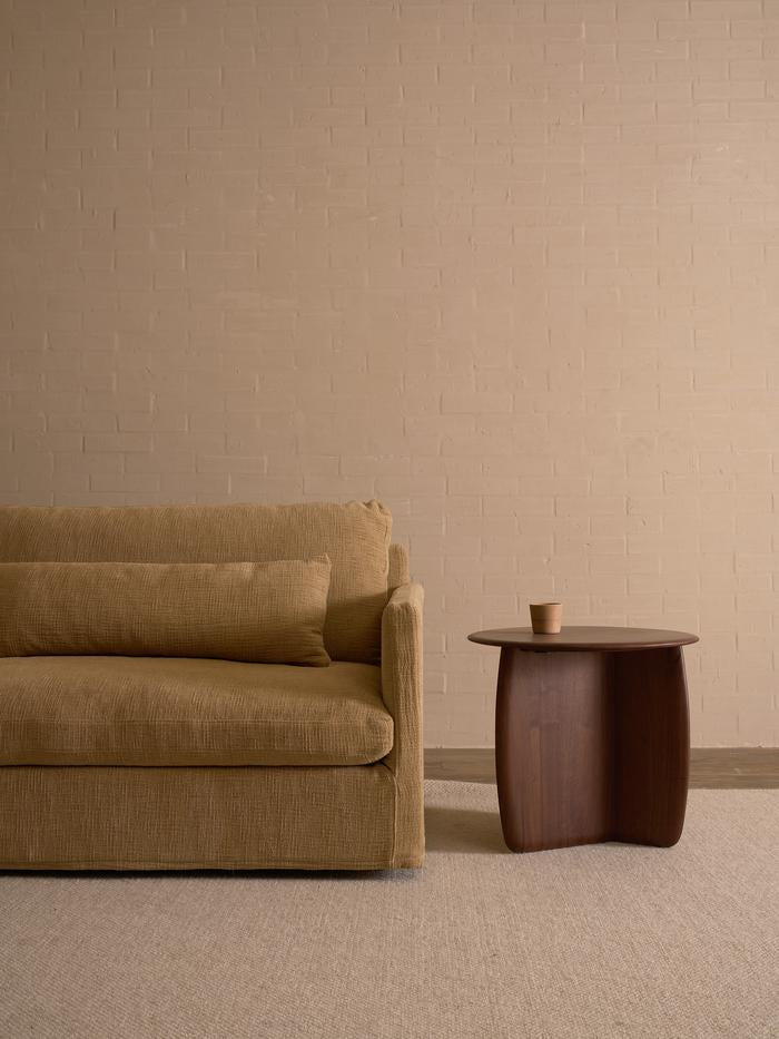 A beige sofa with a matching cushion sits beside the Wooden Stories Eden Side Table in Espresso Oak, which holds a brown cup, set against a plain tan brick wall and a light carpeted floor.