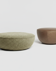 Two round ottomans from the Wooden Stories collection sit on a plain white background: one is a Fable Ottoman in light green fabric, and the other is in brown leather.