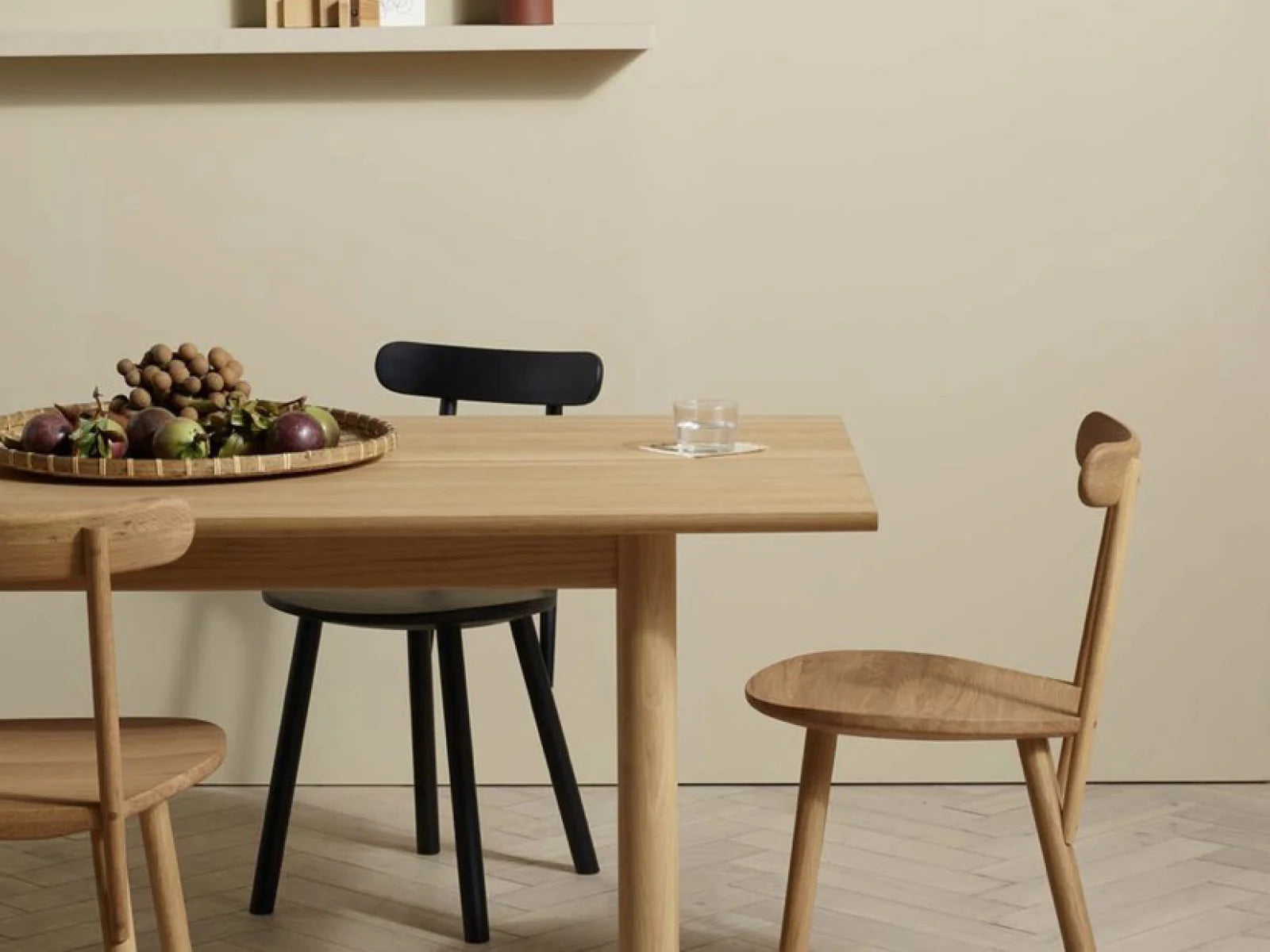 A minimalist dining area includes a light wooden table, a Wooden Stories Figura Dining Chair in Light Oak, two matching chairs, a fruit tray, and a glass of water. A beige wall and floating shelf complete the heritage woodwork atmosphere.