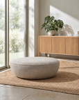 A round, light gray Fable Ottoman | Fabric by Wooden Stories from the Danish Collection sits on a woven rug in a bright room with a large window, a wooden sideboard, and a potted plant on top, offering an inviting extra seat for guests.