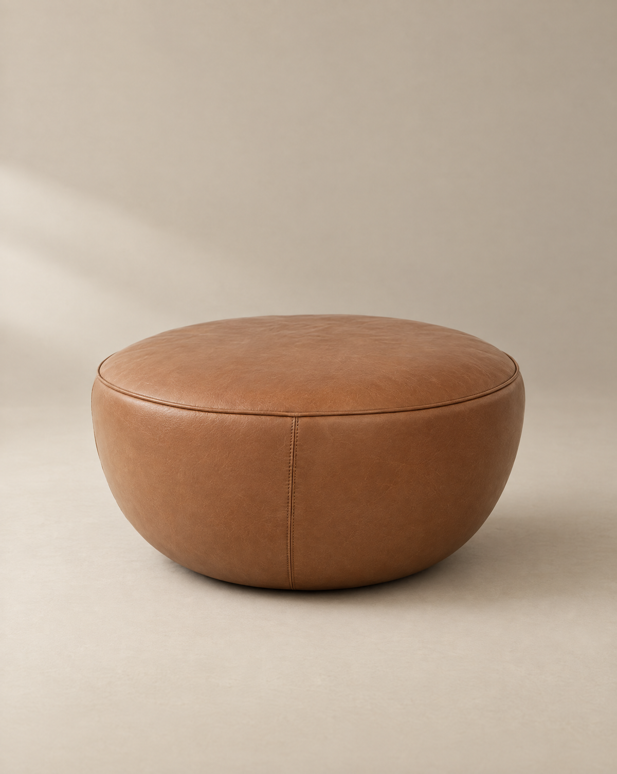 Round, brown Fable Ottoman | Leather by Wooden Stories from the Danish Collection, featuring visible stitching and a smooth, minimalist design. Placed on a neutral background, the ottoman appears soft and comfortable.