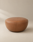 Round, brown Fable Ottoman | Leather by Wooden Stories from the Danish Collection, featuring visible stitching and a smooth, minimalist design. Placed on a neutral background, the ottoman appears soft and comfortable.