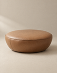 The Fable Ottoman | Leather by Wooden Stories features a round silhouette crafted from brown leather with smooth stitching, showcased on a neutral background to highlight its simple and modern design.