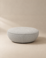 A round, light gray fabric Fable Ottoman by Wooden Stories rests on a beige surface with a matching beige background. Featuring a soft, cushioned look and a clean, modern design, this ottoman is an ideal extra seat for any contemporary setting.