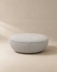 A round, light gray fabric Fable Ottoman by Wooden Stories rests on a beige surface with a matching beige background. Featuring a soft, cushioned look and a clean, modern design, this ottoman is an ideal extra seat for any contemporary setting.