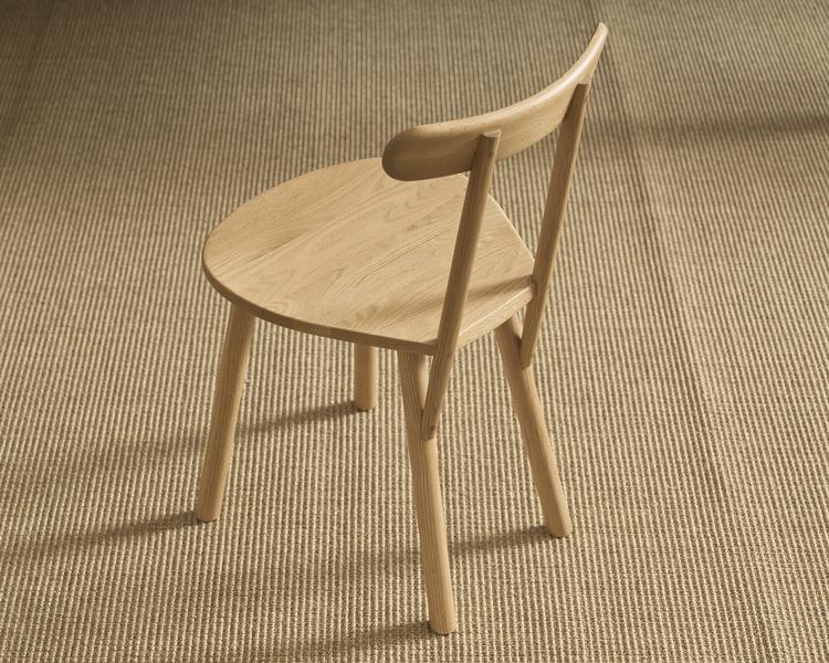 The Figura Dining Chair in Light Oak by Wooden Stories features a curved backrest and three legs, showcasing heritage woodworking with its simple design. It&#39;s seen from above on a textured beige woven rug, highlighting its modern elegance.