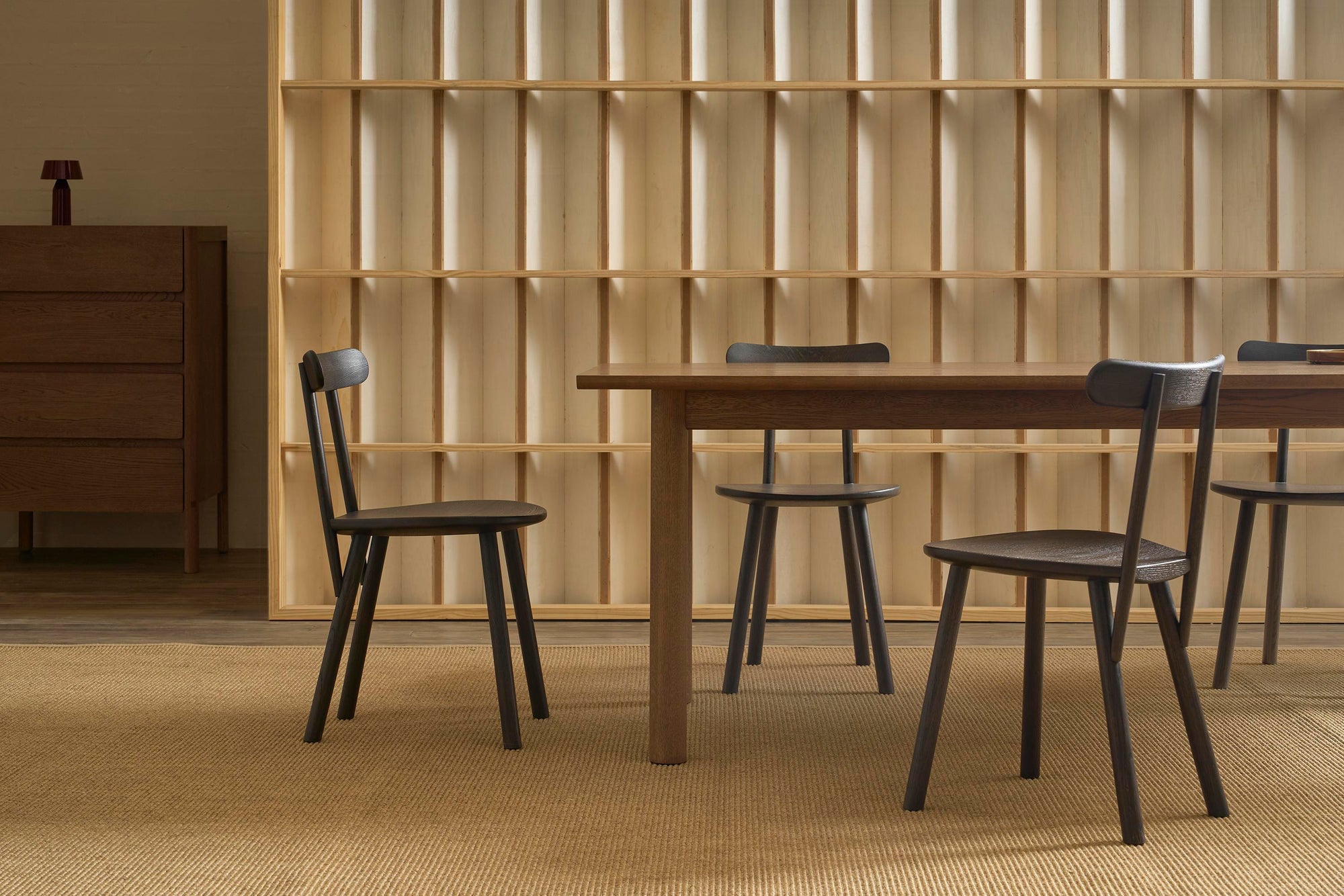 Minimalist dining room featuring a wooden table, one modern milking chair, and three Wooden Stories Figura Dining Chairs in Black Oak, set on a woven rug. A wooden shelving unit and dresser complete the warm, modern look.