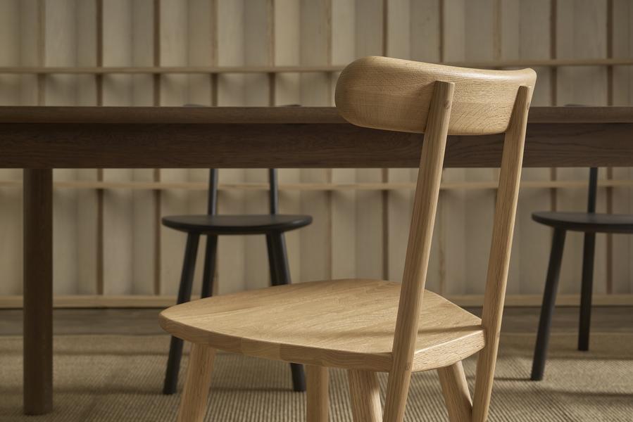 A Figura Dining Chair in Light Oak by Wooden Stories sits before a dark table, with two darker chairs partially visible against a heritage wood-paneled wall.