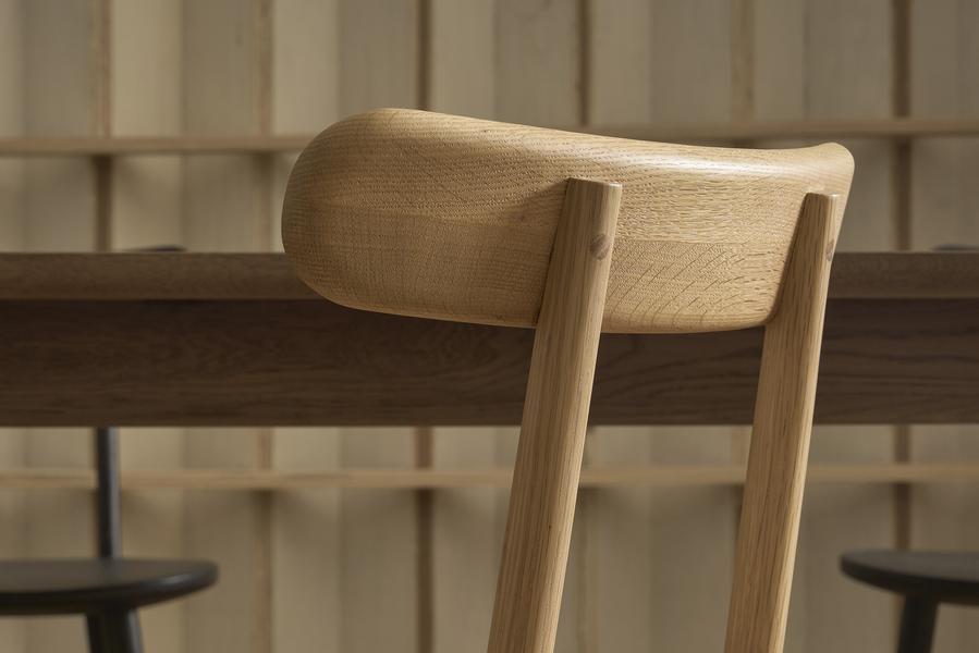 Close-up of the Wooden Stories Figura Dining Chair in Light Oak, featuring a curved backrest and set before a wooden table. Vertical wooden panels and two more chairs complete this timeless heritage-inspired scene.