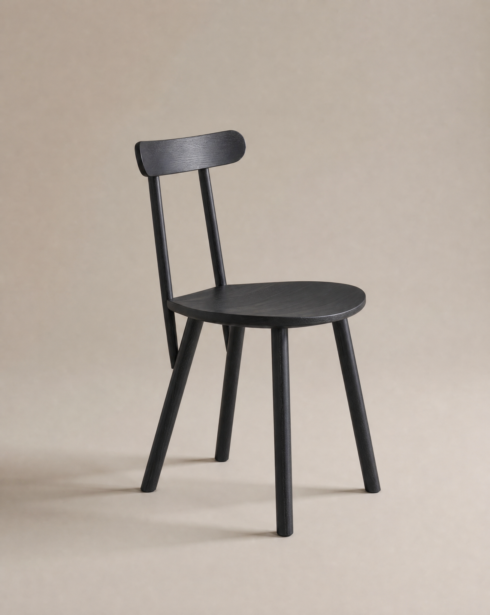 The Figura Dining Chair by Wooden Stories, shown in black oak with a rounded seat, three straight legs, and a curved backrest on a beige background, is ideal for contract-grade spaces and modern interiors.