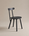 The Figura Dining Chair by Wooden Stories, shown in black oak with a rounded seat, three straight legs, and a curved backrest on a beige background, is ideal for contract-grade spaces and modern interiors.