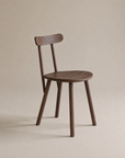 The Figura Dining Chair by Wooden Stories features an espresso oak finish, a round seat, four straight legs, and a curved backrest, shown against a plain beige background.