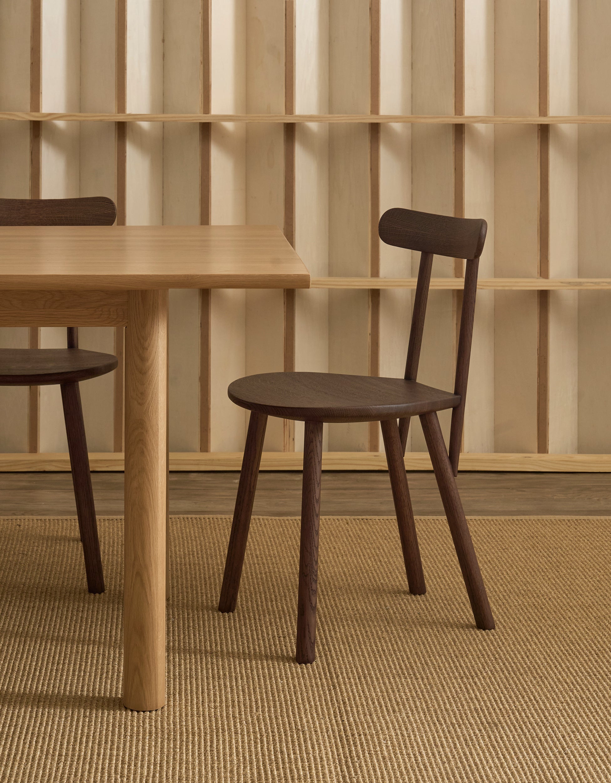A Figura Dining Chair | Espresso Oak by Wooden Stories, featuring dark-stained seat and legs, sits beside a light wood table on a woven rug, with a geometric wooden shelving pattern in the background. This contract-grade chair combines modern Scandinavian style and durability, making it an ideal choice for both residential and commercial spaces.