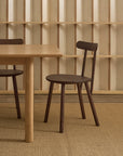 A Figura Dining Chair | Espresso Oak by Wooden Stories, featuring dark-stained seat and legs, sits beside a light wood table on a woven rug, with a geometric wooden shelving pattern in the background. This contract-grade chair combines modern Scandinavian style and durability, making it an ideal choice for both residential and commercial spaces.