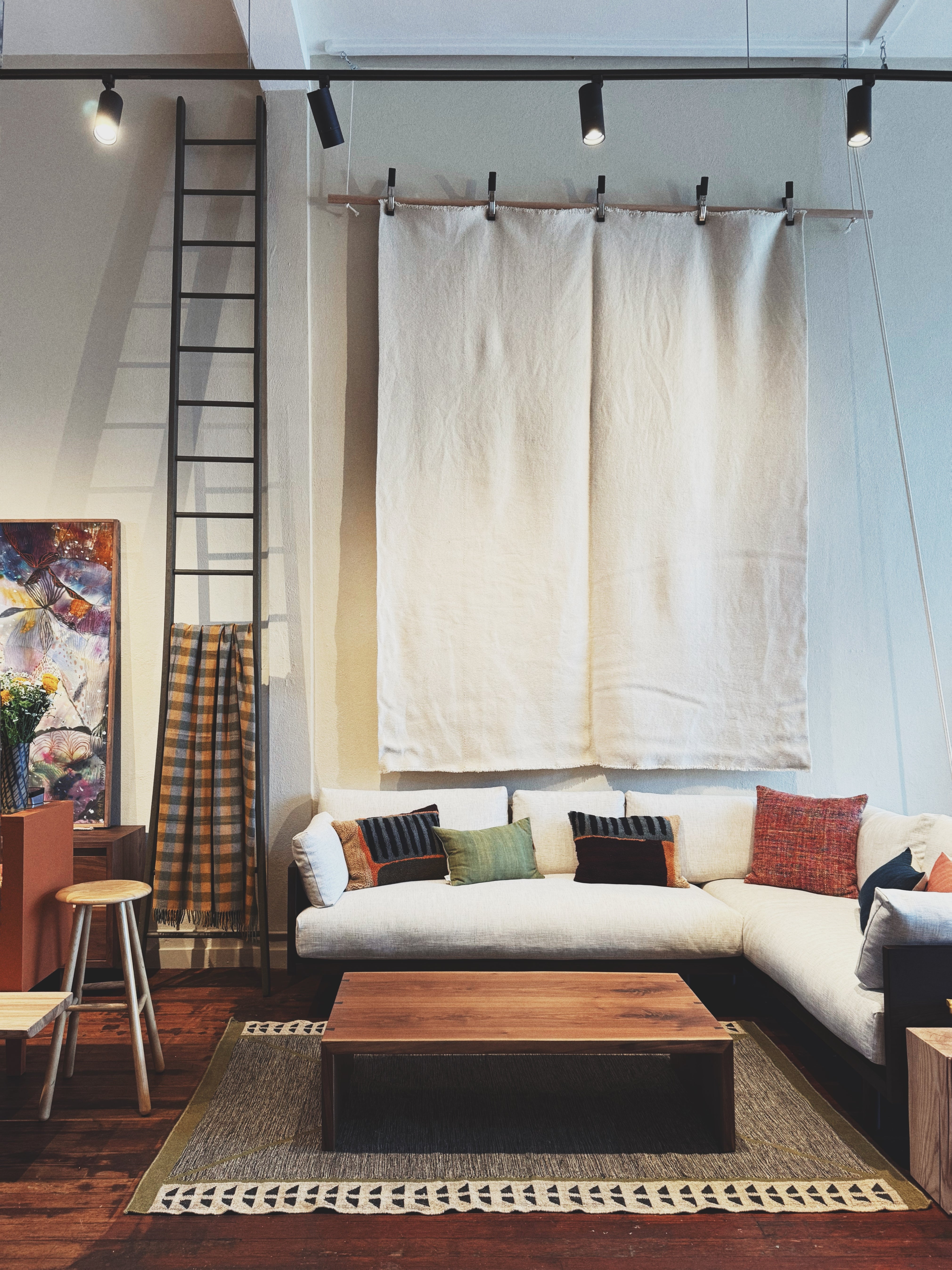 Sobu interior with a low wooden coffee table, sectional sofa with colorful cushions, wall art, a ladder, and a large neutral fabric wall hanging.


