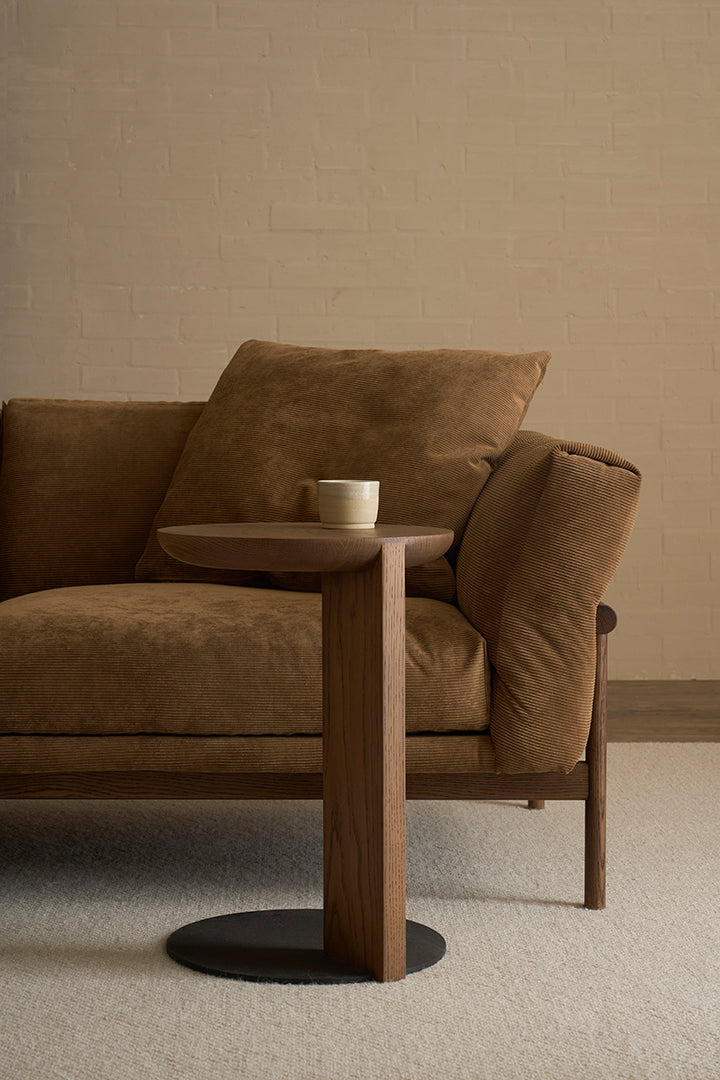 A cozy brown sofa with plush cushions sits beside the Wooden Stories Guild Side Table in Espresso Oak, which holds a white cup, all set against a neutral brick wall and beige carpeted floor.