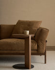 A cozy brown sofa with plush cushions sits beside the Wooden Stories Guild Side Table in Espresso Oak, which holds a white cup, all set against a neutral brick wall and beige carpeted floor.