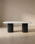 The Tathra Ellipse Dining Table by Wooden Stories features a modern oval stone top and two wide cylindrical black legs, displayed against a neutral beige background.