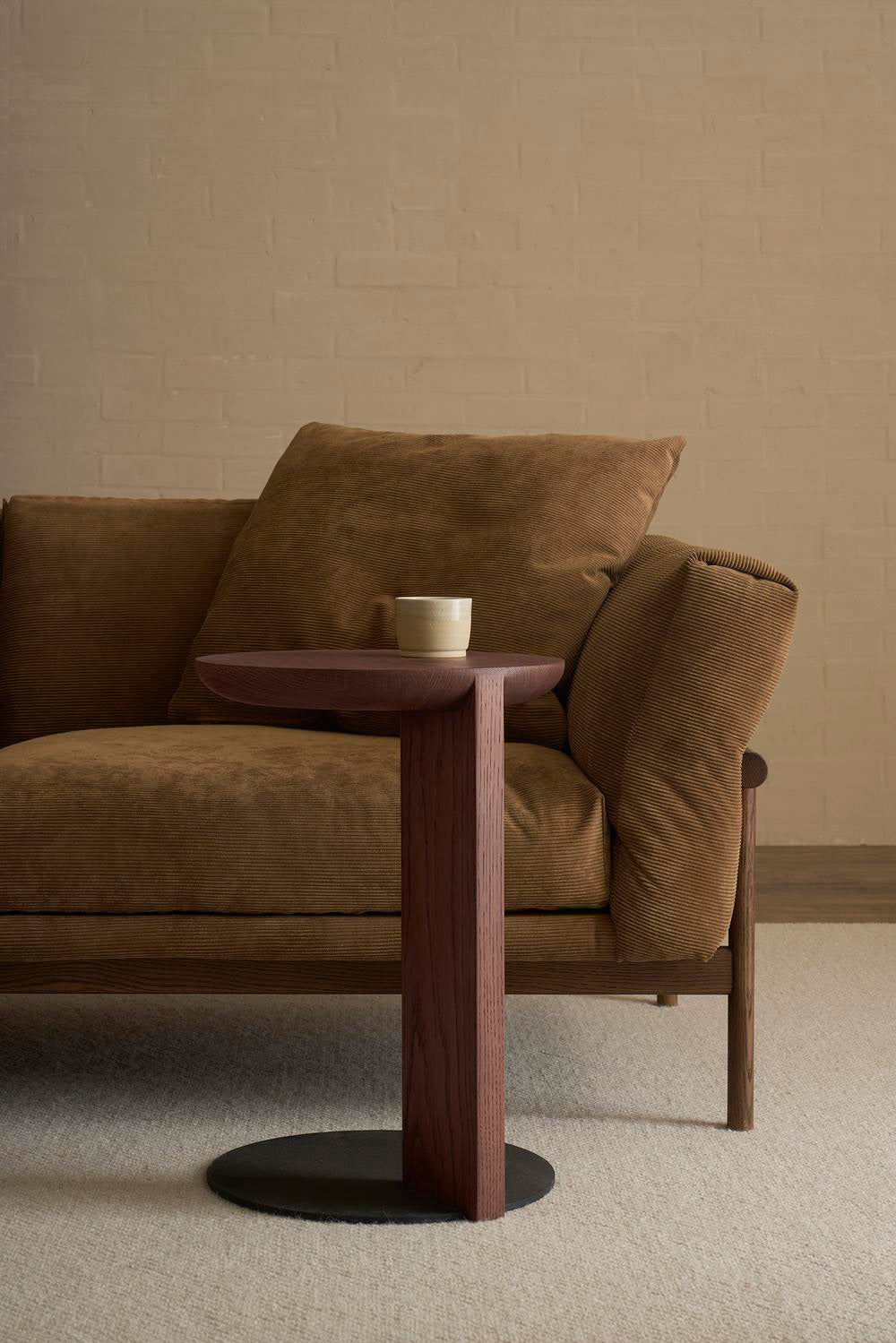 A modern brown sofa with plush cushions stands next to the Wooden Stories Guild Side Table in Espresso Oak, which holds a ceramic cup, set against a light brick wall and carpeted floor in this minimalist contract-grade room.