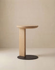 The Guild Side Table by Wooden Stories features a minimalist design with a solid oak round top, single vertical leg, and flat black circular base, shown against a plain beige background.
