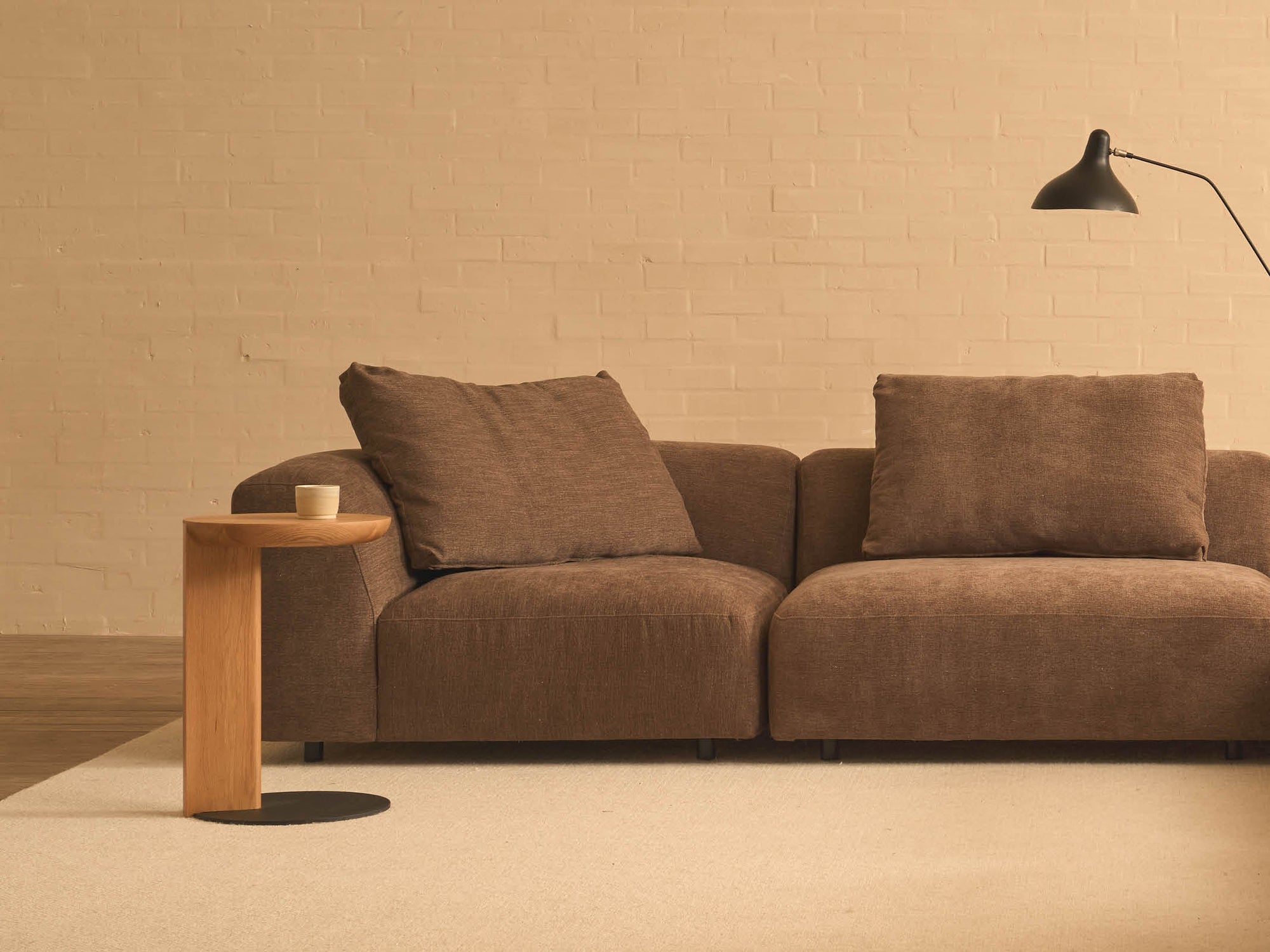 A modern living room with a brown sofa, a beige rug, and a black wall-mounted reading lamp against a light brick wall features the Wooden Stories Guild Side Table in Light Oak holding a cup.