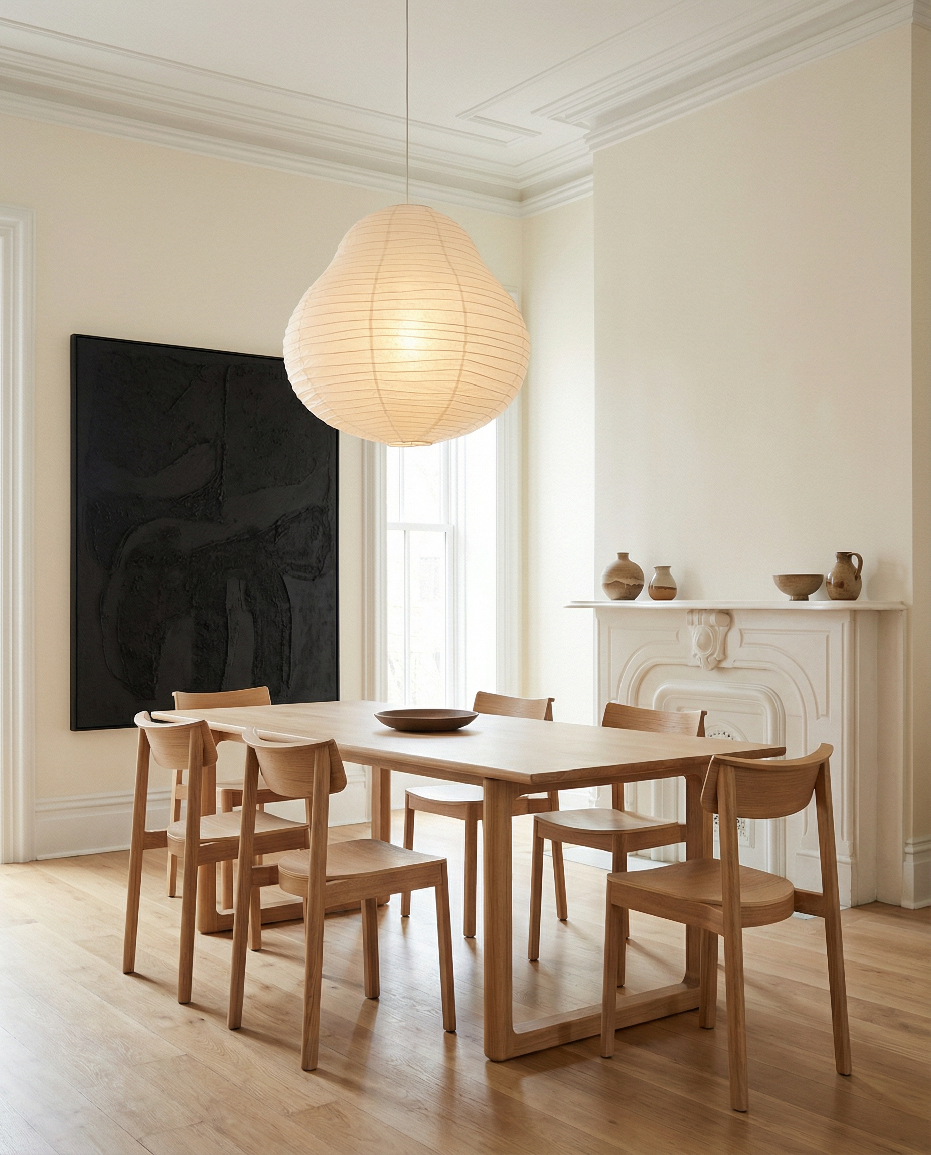 A minimalist dining room featuring the Wooden Stories Hover Dining Table in Light Oak with six matching chairs, a large paper lantern pendant, neutral walls, dark abstract art, and simple pottery on a white mantel.